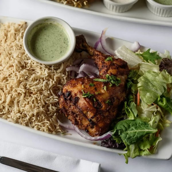 Grilled chicken leg with rice, green sauce, red onions, and a side salad on a white plate.