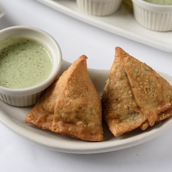Two golden-brown samosas on a white plate with a small bowl of green chutney.