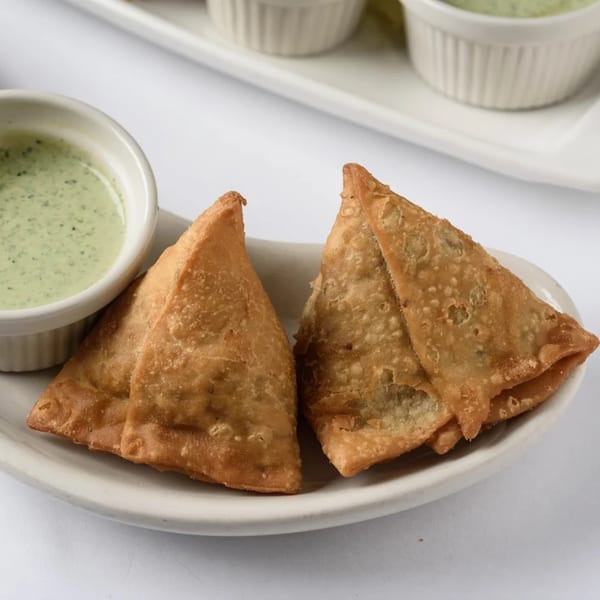 Two golden-brown samosas and a small white bowl of green chutney on a white plate.